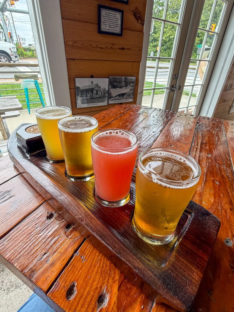 Four colorful craft beers on a wooden flight board inside a cozy brewery.